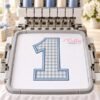 Machine embroidery mockup showing a number 1 applique on fabric in a hoop centered beneath the needles of a multi-needle embroidery machine.