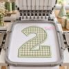 Number 2 applique with sage gingham fabric and satin-stitch edging in a gray hoop centered in a multi-needle embroidery machine, with birthday props nearby.