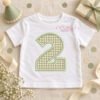 White toddler shirt with a sage gingham number 2 applique and sage satin-stitch border, styled with a birthday hat, wrapped gift, ribbon, and confetti.