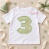 White toddler shirt with a sage gingham number 3 applique, styled on ivory linen with a sage birthday hat, wrapped gift, cream ribbon, and confetti.