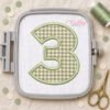 Sage gingham number 3 applique on white fabric in a gray plastic machine embroidery hoop, surrounded by sage thread, gold scissors, and birthday confetti.