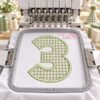Sage gingham number 3 applique on white fabric in a gray hoop centered beneath the raised needles of a multi-needle embroidery machine.
