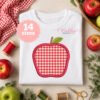 Apple applique embroidery design stitched on a folded white shirt with apples, thread and sewing supplies