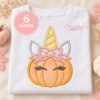 Unicorn pumpkin appliqué mockup on a folded white shirt, featuring peach dotted fabric, a pink floral bow, closed eyelash eyes, and a yellow spiral horn.