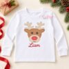 White children’s shirt laid flat with the boy reindeer quick stitch design and the name Liam embroidered below it in red.