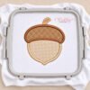Acorn applique embroidery design stitched on white fabric in a plastic machine embroidery hoop