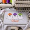 Boy Ghosts with Baseball Hats Quick Stitch Embroidery Design _ Logo 2 Boy ghost trio on a gray shirt in a multi-needle embroidery machine, with orange, purple, and lime hats and Oliver in orange sketch lettering.