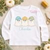Boy Ghosts with Hats Trio Applique Design _ Logo 1 White long sleeve shirt laid flat with three smiling ghost appliques wearing gingham baseball hats and the name Charlie embroidered in aqua below.