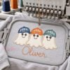 Boy Ghosts with Hats Trio Applique Design _ Logo 3 Mockup of a multineedle embroidery machine stitching a ghost trio applique on a gray sweatshirt, with navy, orange, and teal hats and Oliver in orange below.