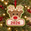 Boy Reindeer Christmas Applique Design _ Logo 3 Reindeer applique ornament with red embroidered 2026 on its cream panel, hanging from a Christmas tree by ribbon through the stitched eyelet.