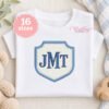 White children's top featuring an illustrative crest applique embroidery design with blue monogram lettering and cream gingham fabric.