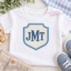 Crest applique monogram embroidery example with cream gingham fabric center, denim blue satin border, and JMT initials.