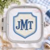 Crest applique embroidery design mockup in a rectangular plastic machine embroidery hoop with blue satin stitching and gingham fabric.
