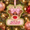 Girl Reindeer Ornament Applique Design _ Logo 1 Girl reindeer applique ornament with a pink bow and pink 2026 lettering, hanging on a Christmas tree by pink ribbon through its green stitched eyelet.