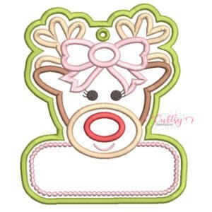 Girl reindeer ornament applique embroidery design with a bow, eyelashes, red nose, stitched hanging eyelet, and blank personalization panel bordered by pink dots.