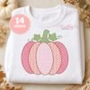 Pink pumpkin with decorative patterned stitching, green leaves, and curling vines displayed on a folded white sweatshirt surrounded by neutral autumn props.