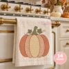 Orange and gold motif pumpkin with green leaves and vines displayed on an ivory kitchen towel hanging from a brass oven handle in an autumn kitchen.