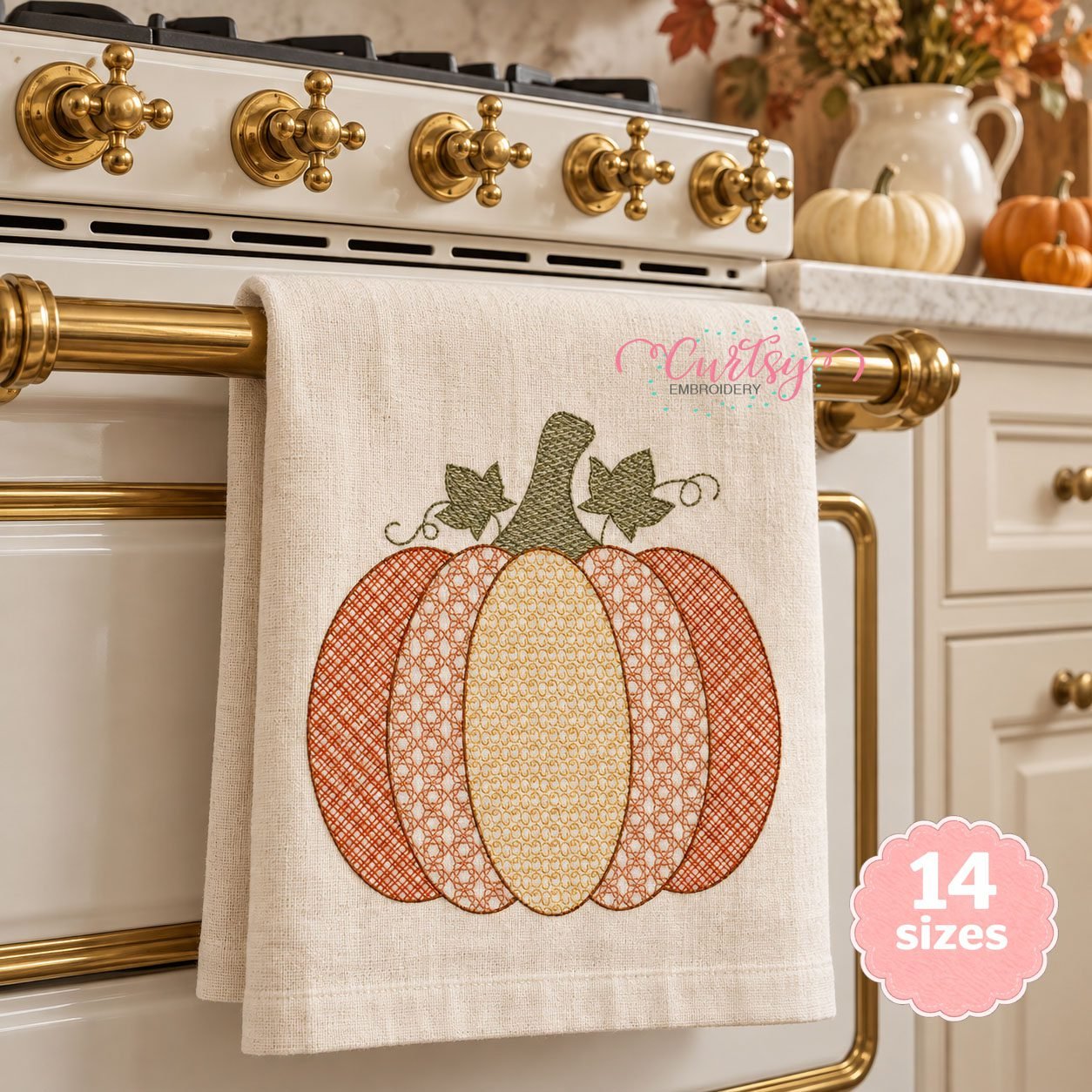 Orange and gold motif pumpkin with green leaves and vines displayed on an ivory kitchen towel hanging from a brass oven handle in an autumn kitchen.