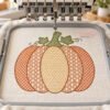 Orange and gold patterned pumpkin embroidery on ivory fabric in a gray hoop beneath a multi-needle embroidery machine, with green leaves and curling vines.