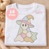 Digital mockup of a folded white shirt featuring a pastel patchwork ghost wearing a witch hat, with decorative embroidery motifs in pink, green, blue, ivory, and gold, surrounded by neutral fall props.