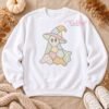 Digital mockup of a white sweatshirt laid flat with a pastel patchwork witch-hat ghost on the chest, styled with pumpkins, dried foliage, a knit blanket, and white sneakers.
