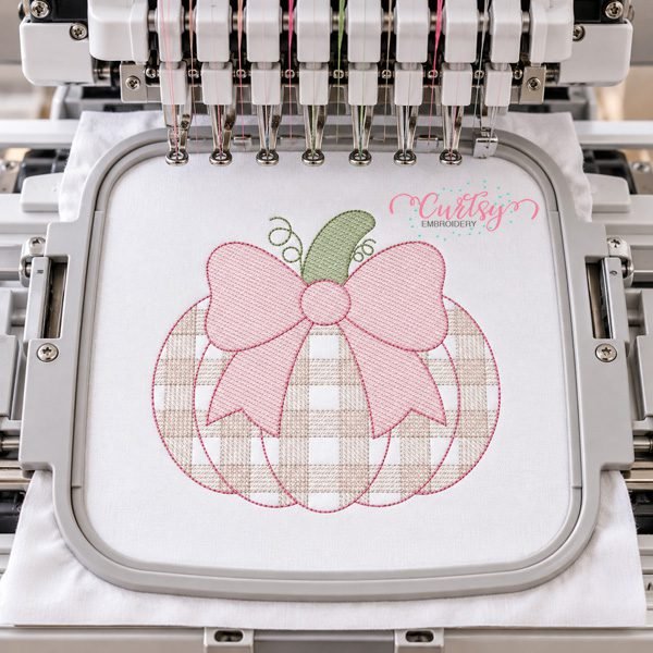 Plaid pumpkin with pink bow embroidery design displayed in a plastic hoop on a multi-needle embroidery machine.