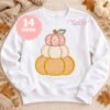 White sweatshirt laid flat with stacked pumpkin applique, styled with autumn leaves, small pumpkins and a neutral knit blanket.