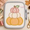 Three stacked applique pumpkins on ivory fabric in a gray plastic machine embroidery hoop, surrounded by coordinating thread spools and scissors.
