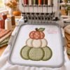 Stacked pumpkin applique shown in a hoop beneath a multi-needle embroidery machine, with satin-stitch pumpkin borders, a stem and a leaf.