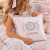 Untitled AI-assisted bedroom lifestyle mockup of a personalized white pillow with a colorful wrapped candy applique, pink gingham ruffle trim, and the embroidered name Emma.